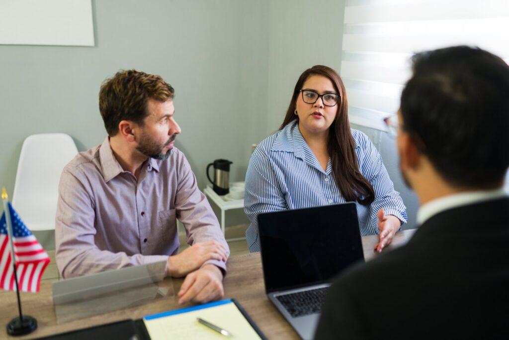Two people sitting at a desk across from another person with a laptop and an American flag on the table